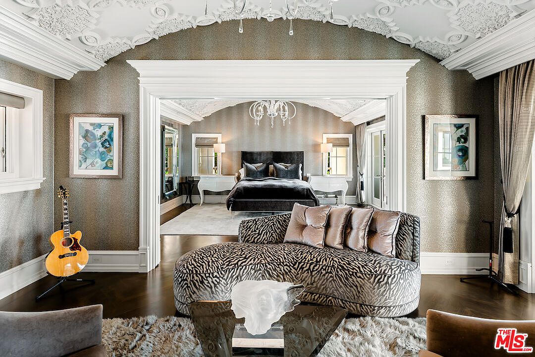 This is a luxurious primary bedroom featuring a sitting area with a zebra-print chaise lounge and a glass coffee table. The bedroom is accessed through an arched doorway, leading to a bed with a dark headboard and matching bedding. The walls are covered in a textured wallpaper, and the ceiling has ornate molding, creating an elegant and sophisticated atmosphere.