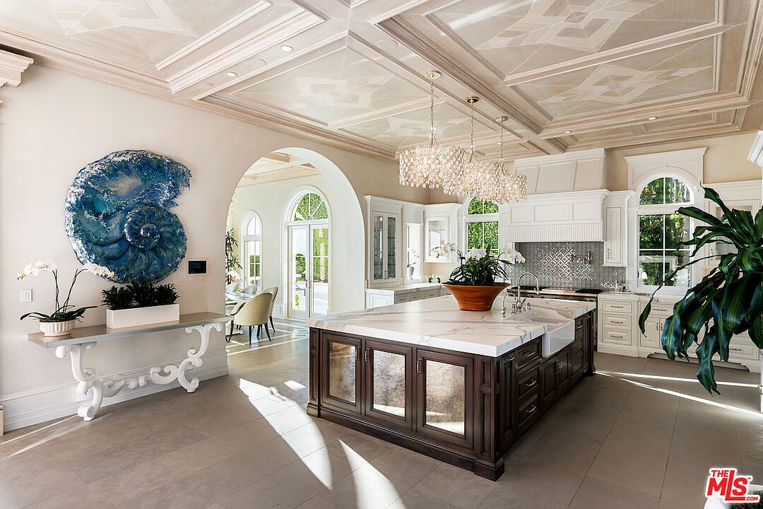This is a bright and luxurious kitchen featuring a large central island with a marble countertop and dark wood cabinetry. The kitchen is well-lit with natural light from arched windows and elegant chandeliers. A decorative blue seashell art piece adds a unique touch to the adjacent wall, complementing the overall high-end design.