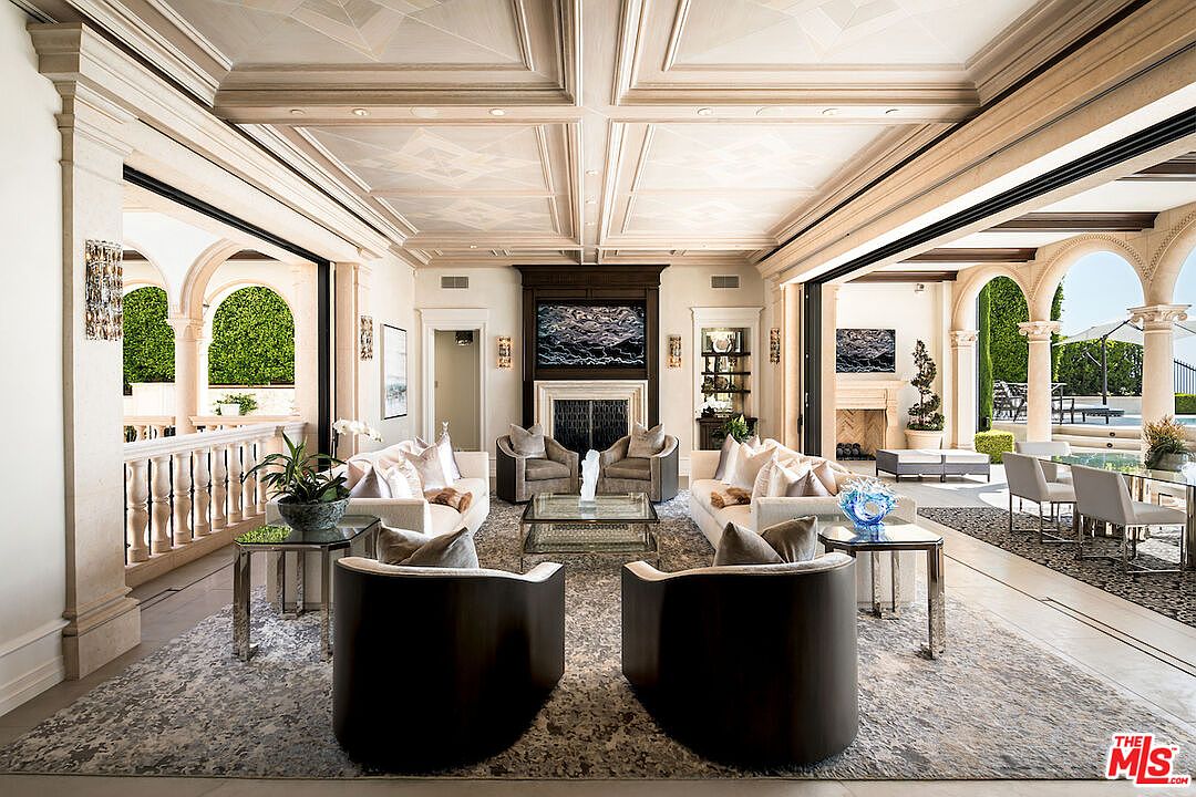 This is a luxurious living room featuring a symmetrical layout with two sofas, four armchairs, and a central coffee table. The room has a coffered ceiling with decorative patterns, a fireplace with a dark wood mantel, and large openings to an outdoor patio area. The overall impression is one of elegance and spaciousness, perfect for entertaining.