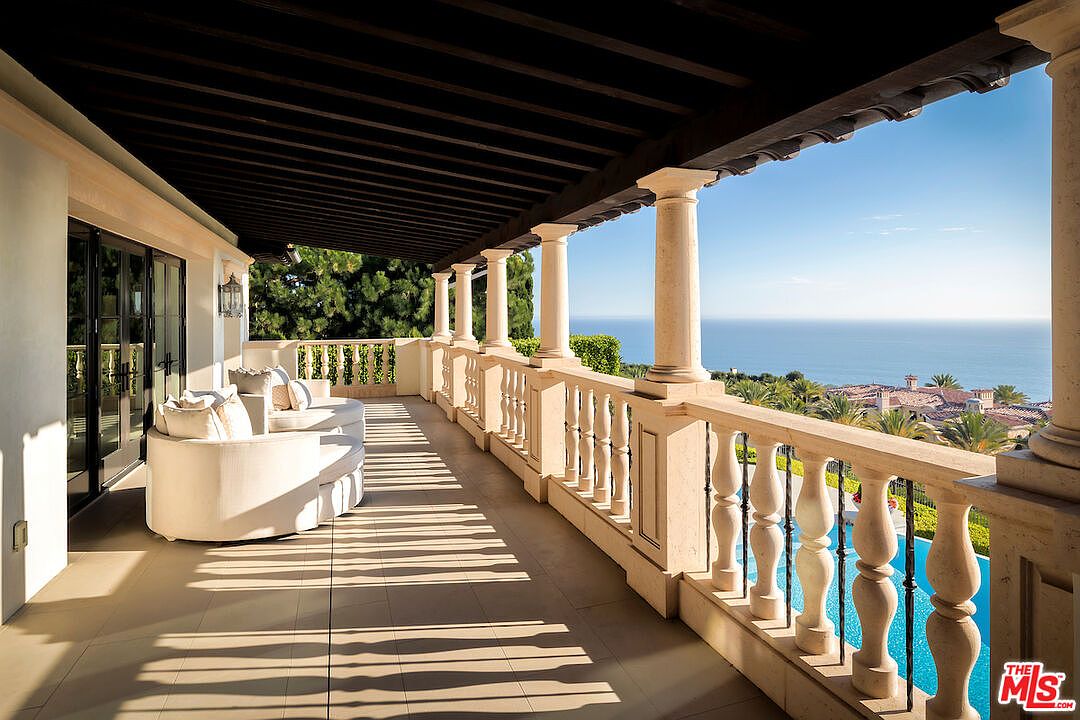 This image showcases a luxurious outdoor balcony or patio area with stunning ocean views. The space features a comfortable, curved sofa with plush cushions, perfect for relaxation. Architectural details include elegant columns, a decorative balustrade, and a dark wood pergola, creating a sophisticated and inviting atmosphere.