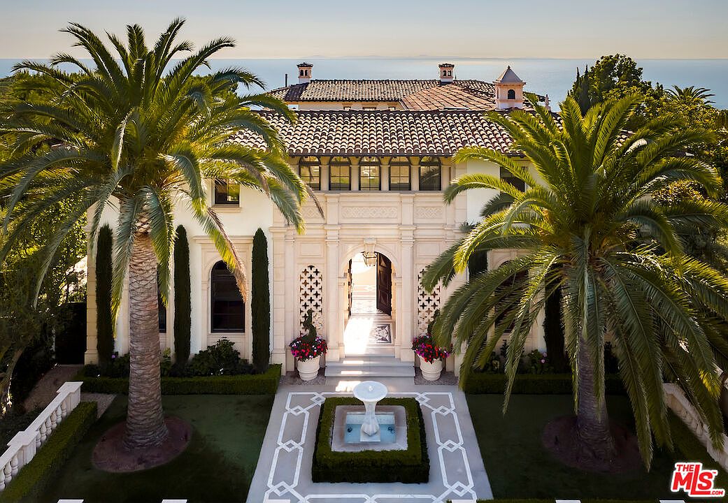 This is a stunning front view of a luxurious estate, showcasing its grand entrance with ornate architectural details, including arched windows and decorative stonework. The property is framed by mature palm trees and meticulously manicured landscaping, leading to a central fountain feature. The overall impression is one of elegance and sophistication, with a glimpse of the ocean in the background adding to the allure.