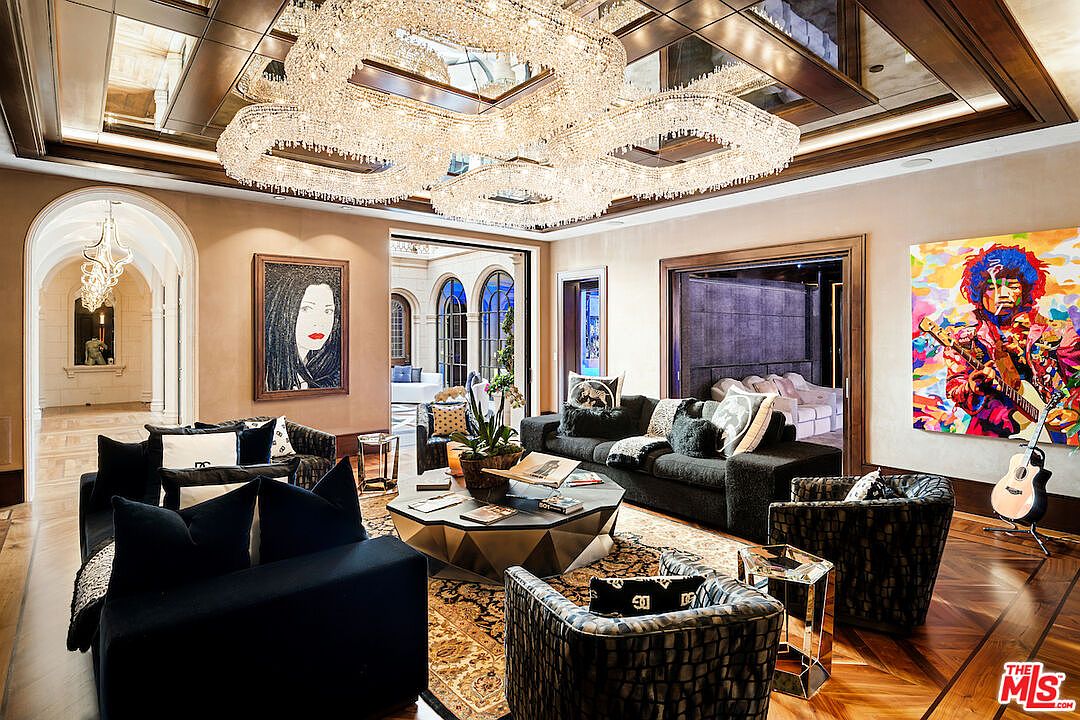This is a luxurious living room featuring a large crystal chandelier, elegant furnishings, and hardwood floors. The room is decorated with artwork, including a portrait and a colorful painting of Jimi Hendrix. The space is open and inviting, with access to an outdoor area through arched doorways, creating a seamless indoor-outdoor flow.