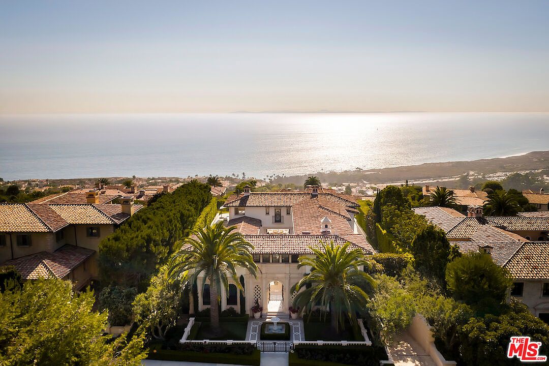 This aerial shot showcases a luxurious estate with a terracotta tile roof, surrounded by lush landscaping and palm trees. The property features a grand entrance with an arched gateway and a meticulously designed garden with a fountain. The backdrop is a stunning ocean view, enhancing the property's appeal and exclusivity.