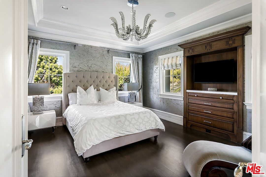 This is a primary bedroom featuring a tufted headboard bed with white bedding, flanked by matching nightstands and lamps. The room has a built-in entertainment center with a dark wood finish and a television. The walls are covered in a textured wallpaper, and the room is illuminated by a modern chandelier.
