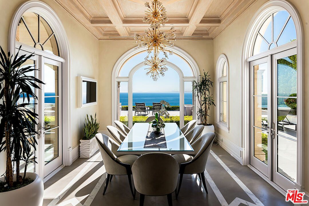 This is an elegant dining room featuring a glass-top table surrounded by upholstered chairs. The room is bathed in natural light from large arched windows offering ocean views. A modern chandelier hangs above the table, and the flooring is a light-colored stone with geometric patterns.