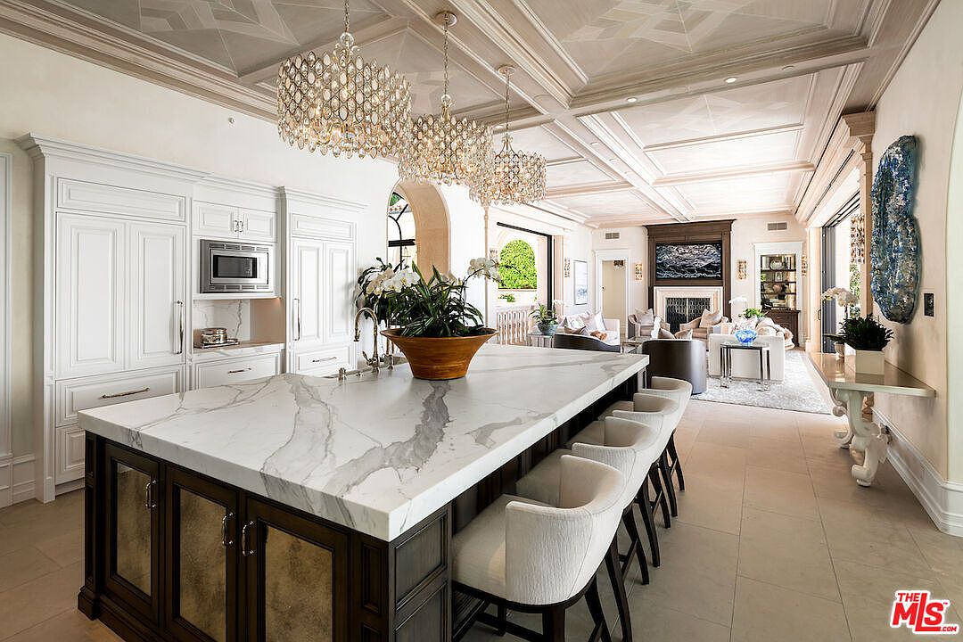 This is a luxurious kitchen featuring a large island with a marble countertop and seating, custom white cabinetry, and high-end appliances. Three crystal chandeliers hang above the island, adding a touch of elegance. The kitchen seamlessly flows into a living area, creating an open and inviting space.