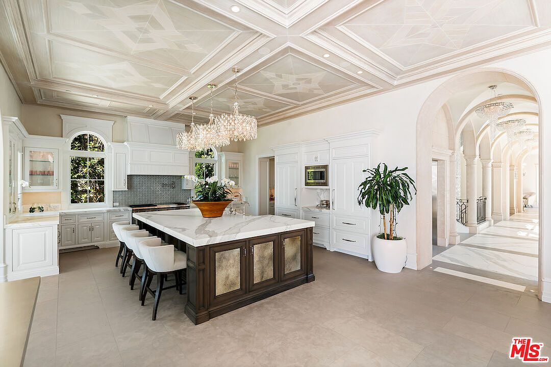 This is a spacious, luxurious kitchen featuring a large central island with a marble countertop and seating. The kitchen is adorned with white cabinetry, stainless steel appliances, and elegant chandeliers. An arched hallway leads to another area, enhancing the grandeur of the space.