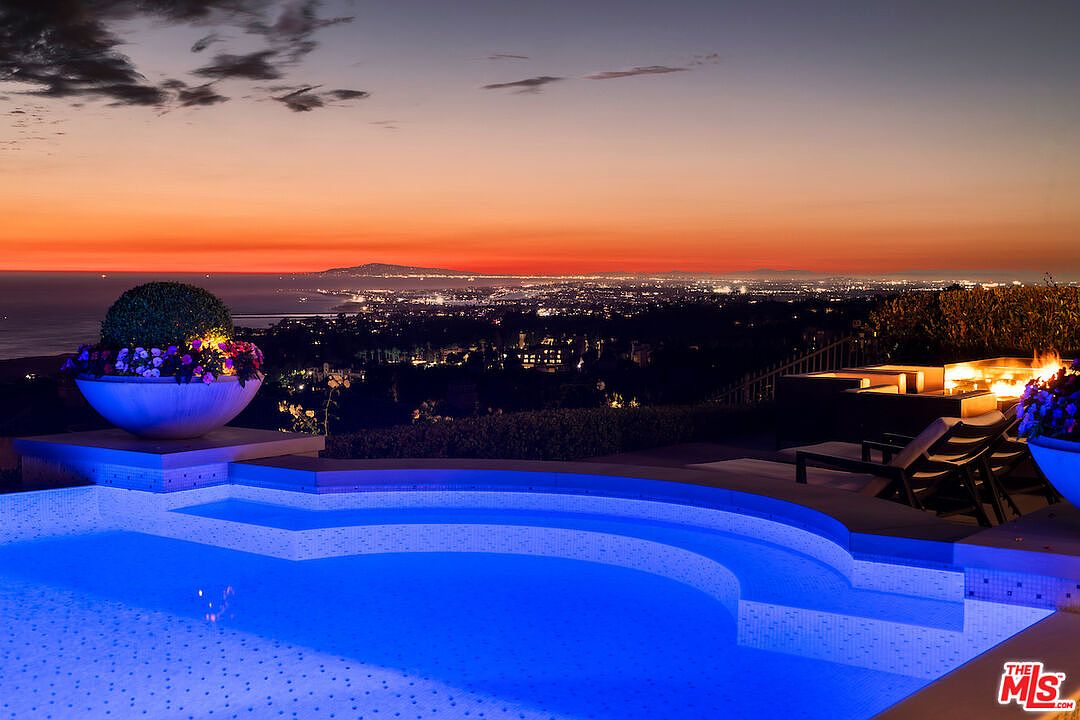 This image showcases a luxurious pool area at dusk, featuring a curved pool with illuminated blue water. The scene includes outdoor lounge furniture, a fire pit, and decorative planters, all set against a backdrop of city lights and a vibrant sunset. The overall impression is one of upscale relaxation and sophisticated outdoor living.