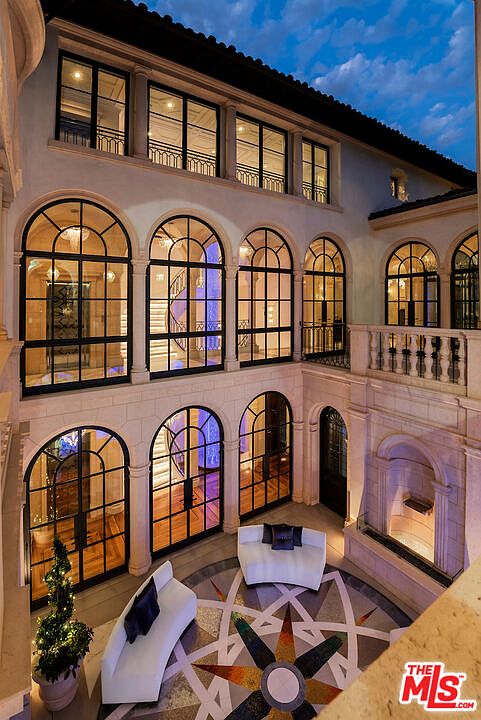 This image showcases a luxurious outdoor patio area, featuring elegant arched windows and doors that provide ample natural light. The patio is furnished with modern white seating arrangements, set against a decorative tile floor with a compass design. The overall impression is one of sophistication and refined outdoor living.