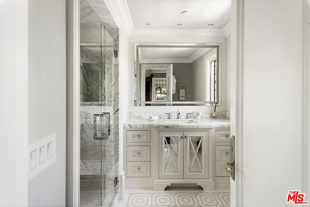 This is a well-lit primary bathroom featuring a marble-topped vanity with mirrored cabinet doors and drawers. A large, framed mirror hangs above the vanity, reflecting the room's neutral color palette. A glass-enclosed shower with marble walls is visible to the left, adding a touch of luxury.