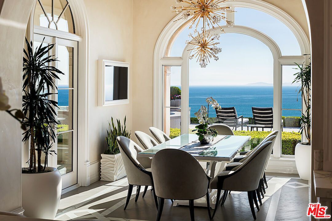 This is an interior shot of a dining room featuring a glass-top table surrounded by upholstered chairs with dark wood legs. Large arched windows offer a stunning ocean view, and a modern chandelier hangs above the table. The room is bright and airy, with a neutral color palette and elegant decor.