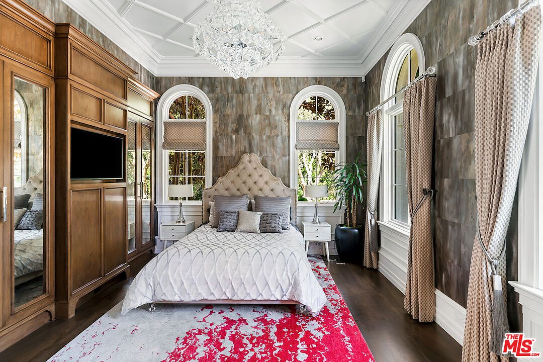 This is a luxurious primary bedroom featuring a tufted headboard bed with decorative pillows, flanked by matching nightstands and lamps. A large wooden wardrobe with a built-in television stands against one wall, while arched windows with custom blinds and curtains provide natural light. The room is decorated with a modern chandelier and a bold red and gray rug, creating an elegant and inviting atmosphere.