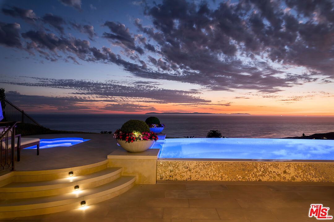 This image showcases a luxurious infinity pool overlooking the ocean at sunset. The pool features integrated lighting, and the surrounding patio includes elegant planters and steps with subtle lighting. The overall impression is one of high-end relaxation and stunning views, perfect for a luxury real estate listing.