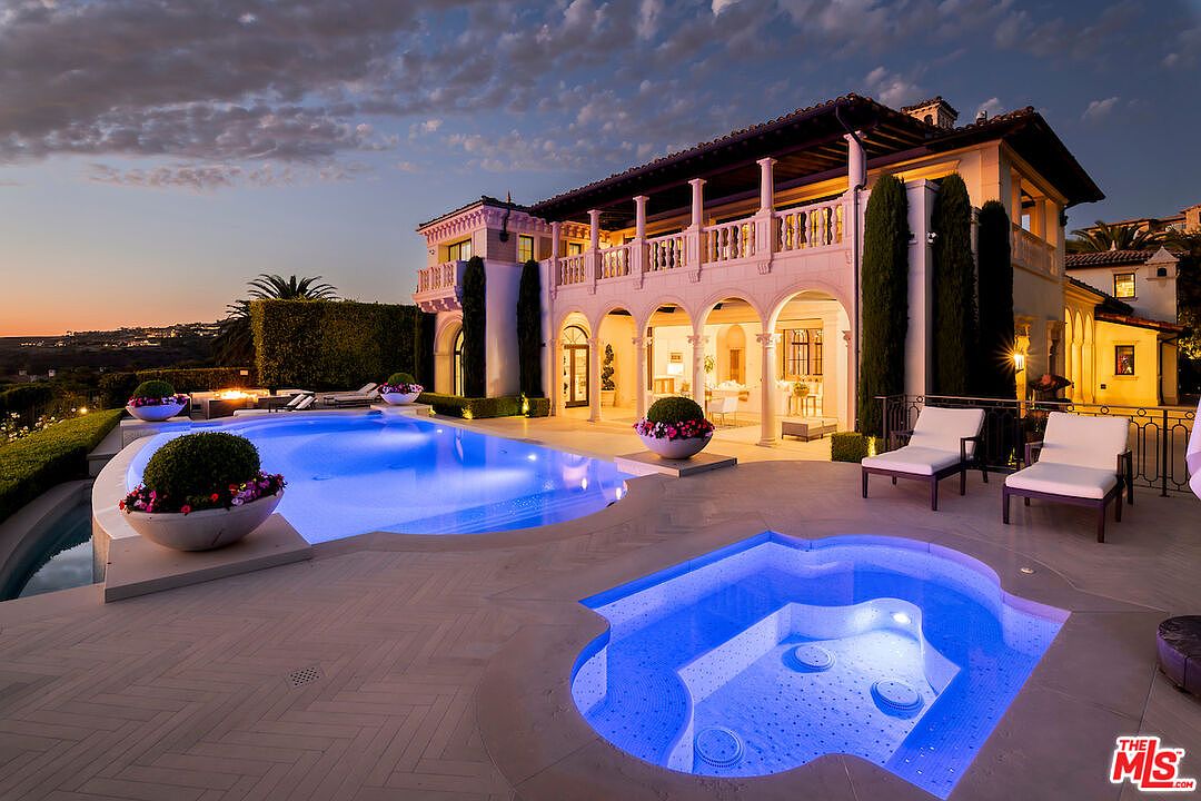 This image showcases a luxurious outdoor pool and spa area at dusk, featuring a large, illuminated pool and a separate, smaller spa with blue lighting. The property boasts a grand Mediterranean-style villa with arched walkways, columns, and a balcony overlooking the pool area. The scene exudes opulence and relaxation, perfect for upscale living and entertaining.