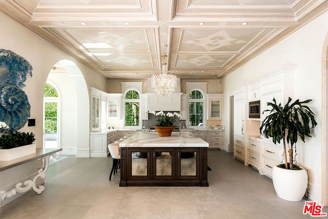 This is a spacious, luxurious kitchen featuring custom white cabinetry, a large marble-topped island with dark wood accents, and a decorative coffered ceiling with a crystal chandelier. Arched windows provide natural light, and a potted plant adds a touch of greenery. The overall impression is one of elegance and high-end design.