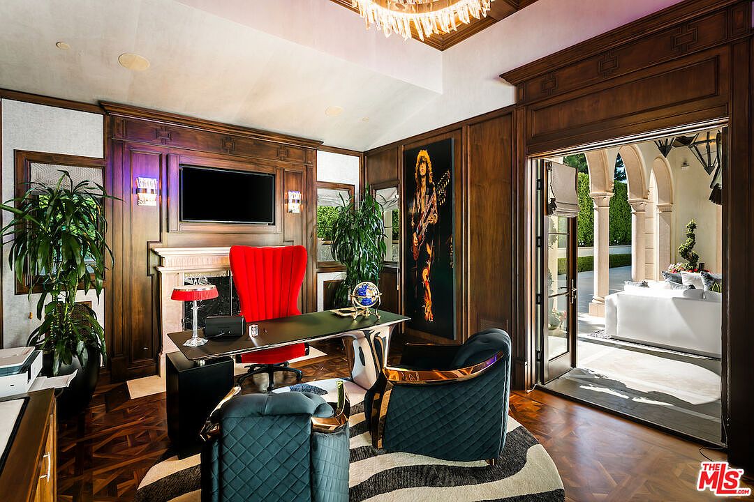This is a luxurious office space featuring dark wood paneling, a large desk with a red high-back chair, and additional seating with blue quilted chairs. A zebra-striped rug adds a modern touch, while a large painting and a flat-screen TV enhance the room's sophistication. The open doorway leads to an outdoor patio, creating a seamless indoor-outdoor flow.