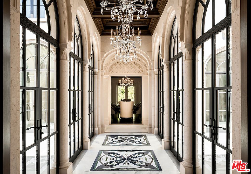 This is an interior shot of a grand hallway featuring arched doorways and windows with black frames. The floor is marble with decorative tile inlays. Two crystal chandeliers hang from the coffered ceiling, adding to the luxurious feel of the space. The perspective is a straight-on view, emphasizing the depth and length of the hallway.