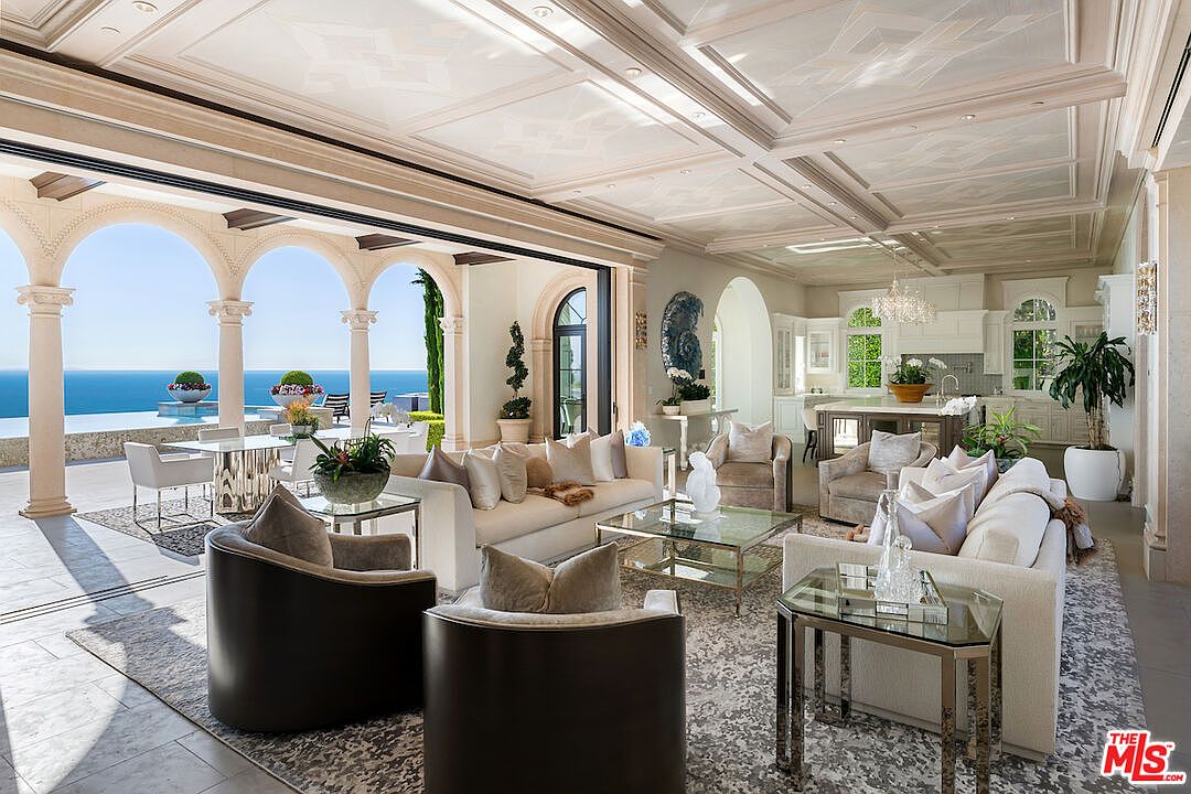 This is a luxurious living room that seamlessly blends indoor and outdoor spaces. The room features elegant furnishings, including plush sofas and armchairs, complemented by glass-topped tables. Large arched openings provide stunning ocean views and access to an outdoor patio, enhancing the room's appeal and creating a serene atmosphere.