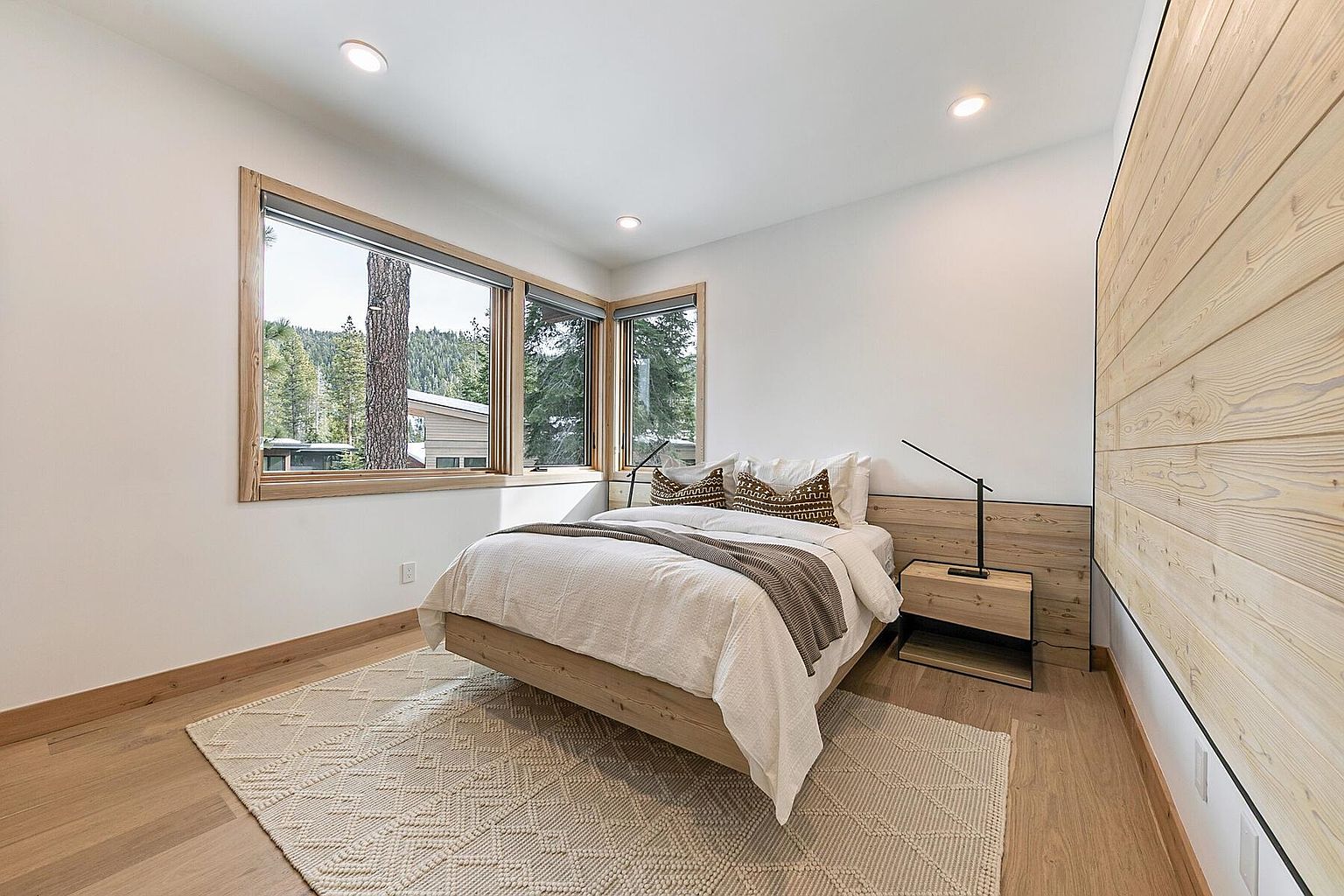 This is a cozy guest bedroom featuring a platform bed with white linens and decorative pillows. A wooden nightstand with a modern lamp sits beside the bed, and a textured rug adds warmth to the hardwood floor. Large windows offer a view of the surrounding trees, creating a serene and inviting atmosphere.