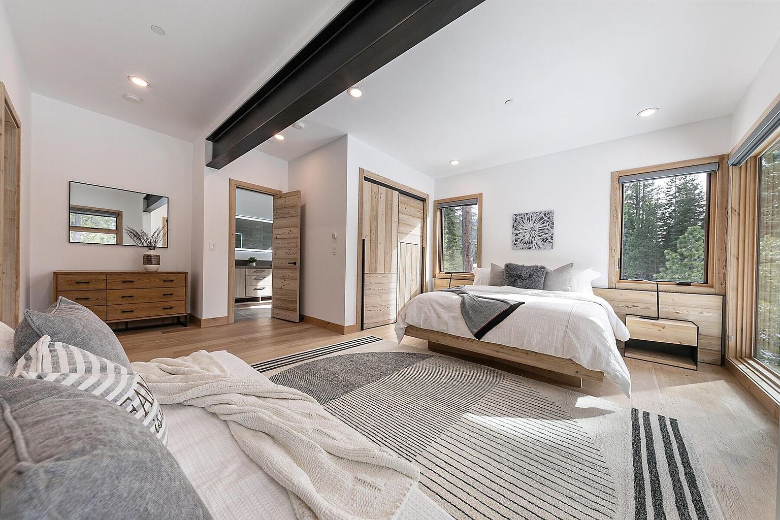 This is a bright and airy primary bedroom featuring a modern design with natural wood accents. The room includes a large bed with white linens, a stylish rug, and a wooden dresser with a mirror. Natural light floods the space through large windows, creating a serene and inviting atmosphere.