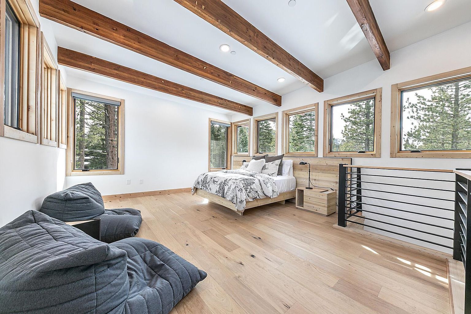This is a bright and airy primary bedroom featuring exposed wooden beams and large windows that offer scenic views of the surrounding trees. The room is furnished with a wooden platform bed, matching nightstands, and comfortable-looking beanbag chairs. The light wood flooring and white walls create a serene and inviting atmosphere, perfect for relaxation.