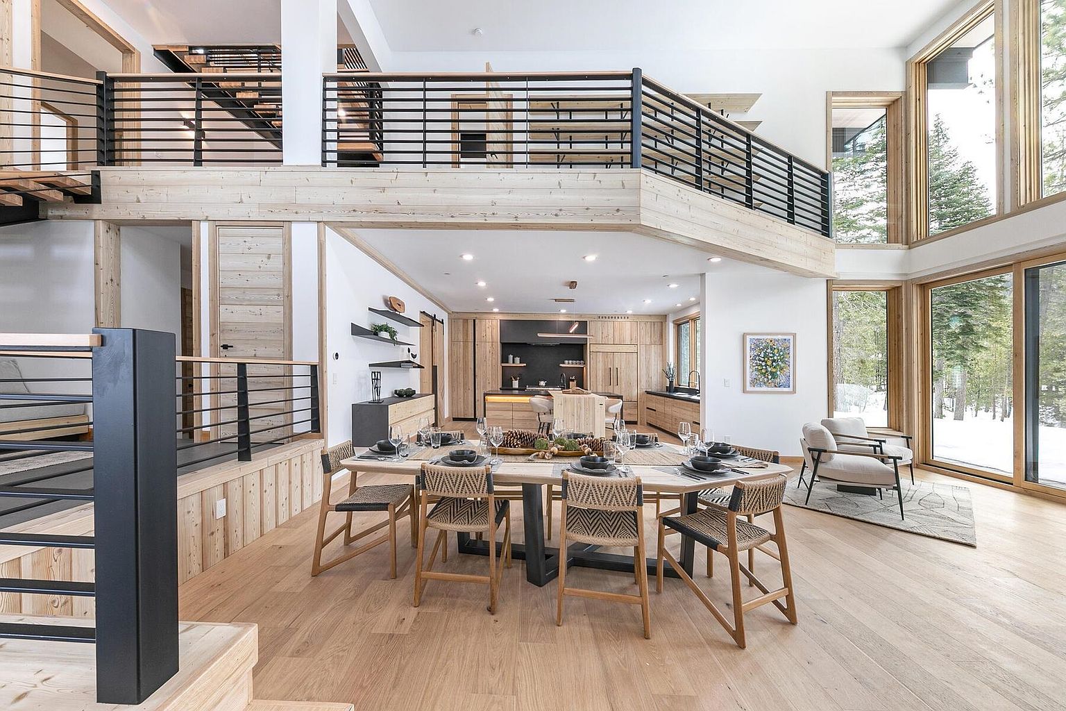 This interior shot showcases a modern dining room with an open-concept design that flows into the kitchen. A large wooden dining table with woven chairs is the focal point, complemented by the natural light streaming through expansive windows. The space features a contemporary aesthetic with wood accents, black metal railings, and a view of a snowy landscape, creating a warm and inviting atmosphere.