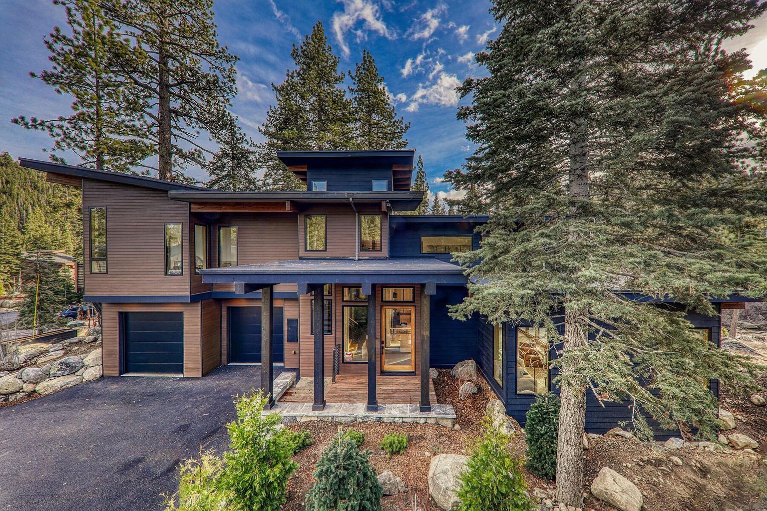 This is a front exterior view of a modern, two-story home with a unique architectural design. The house features a combination of brown and dark blue siding, a covered entryway with dark wooden pillars, and a black garage door. The landscaping includes a mix of greenery and rocks, complementing the natural surroundings and creating a welcoming curb appeal.