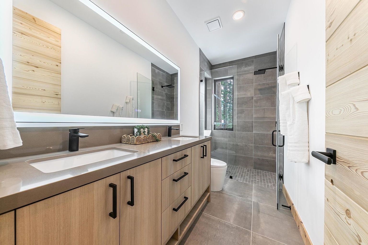 This is a modern primary bathroom featuring a double vanity with light wood cabinetry, black hardware, and a light gray countertop. A large, illuminated mirror hangs above the vanity, reflecting the walk-in shower with gray tiling. The bathroom has a clean, contemporary design with wood accents on the door and wall.