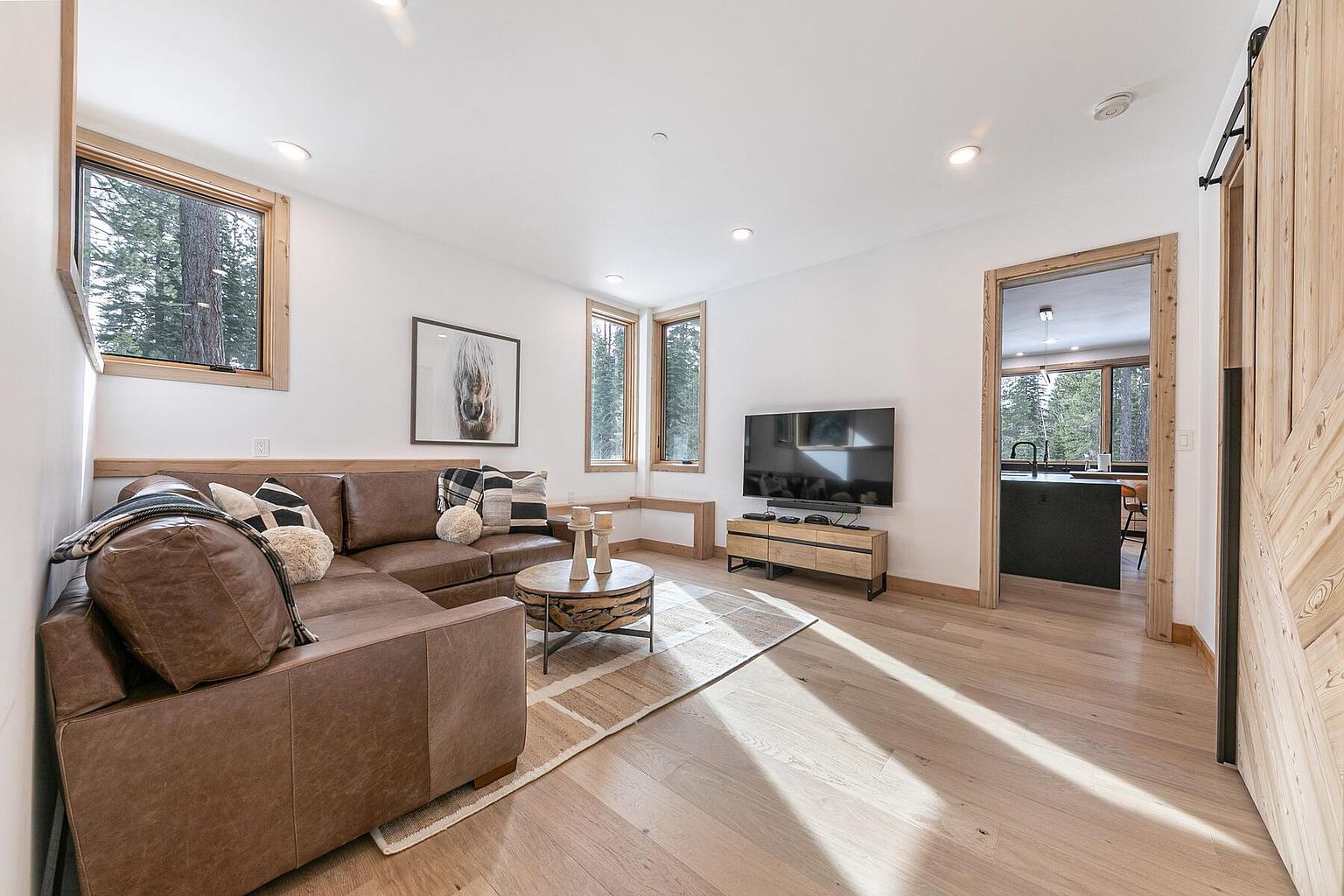 This is an inviting living room featuring a brown leather sectional sofa adorned with decorative pillows and a throw blanket. A round coffee table sits atop a neutral-toned area rug, and a modern entertainment center houses a flat-screen television. Natural light floods the room through multiple windows, highlighting the hardwood flooring and creating a warm, welcoming atmosphere.