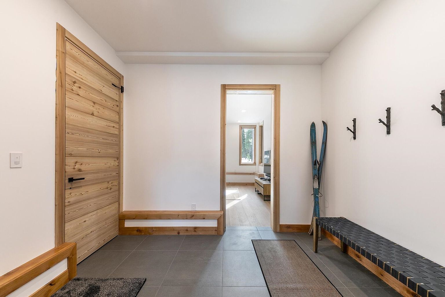 This interior shot showcases a modern hallway with a clean and inviting aesthetic. The hallway features a light wood door, a doorway leading to another room, and a bench with skis leaning against the wall. The flooring is a dark tile, and the walls are painted a bright white, creating a spacious and airy feel.