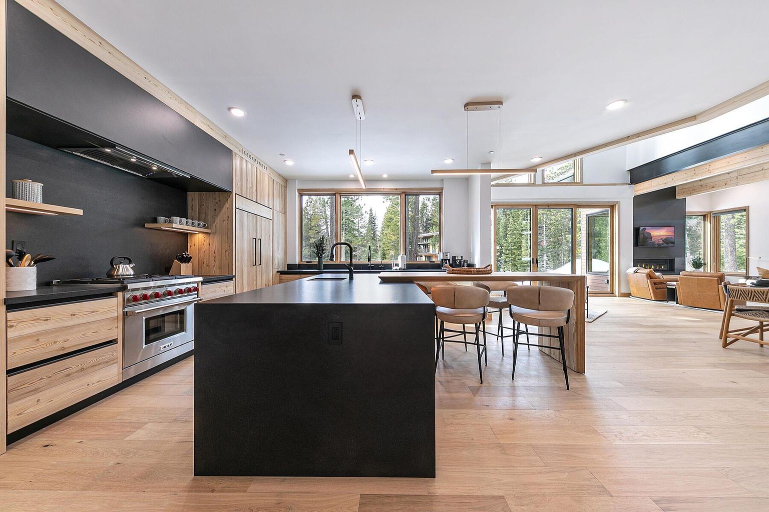 This is a modern kitchen with a large black island, light wood cabinetry, and black countertops. Stainless steel appliances are visible, including a range and hood. Large windows provide natural light and views of the surrounding trees, creating a bright and inviting space that seamlessly blends with the adjacent living area.