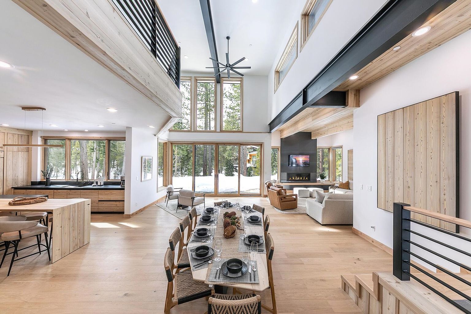 This is an interior shot of a modern dining room and living space. The dining area features a long wooden table set for a meal, surrounded by woven chairs. The living area includes comfortable seating around a fireplace with a television above it. Large windows offer views of a snowy landscape, and the open floor plan creates a spacious and inviting atmosphere.