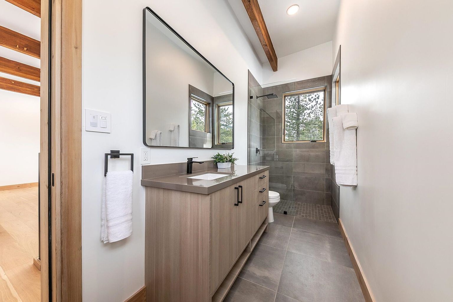 This is a modern bathroom featuring a sleek vanity with a light wood finish and a gray countertop, complemented by a large rectangular mirror with a black frame. The shower area is tiled in gray and enclosed with glass, while the floor is covered in gray tiles. The room is well-lit and has a clean, contemporary aesthetic.