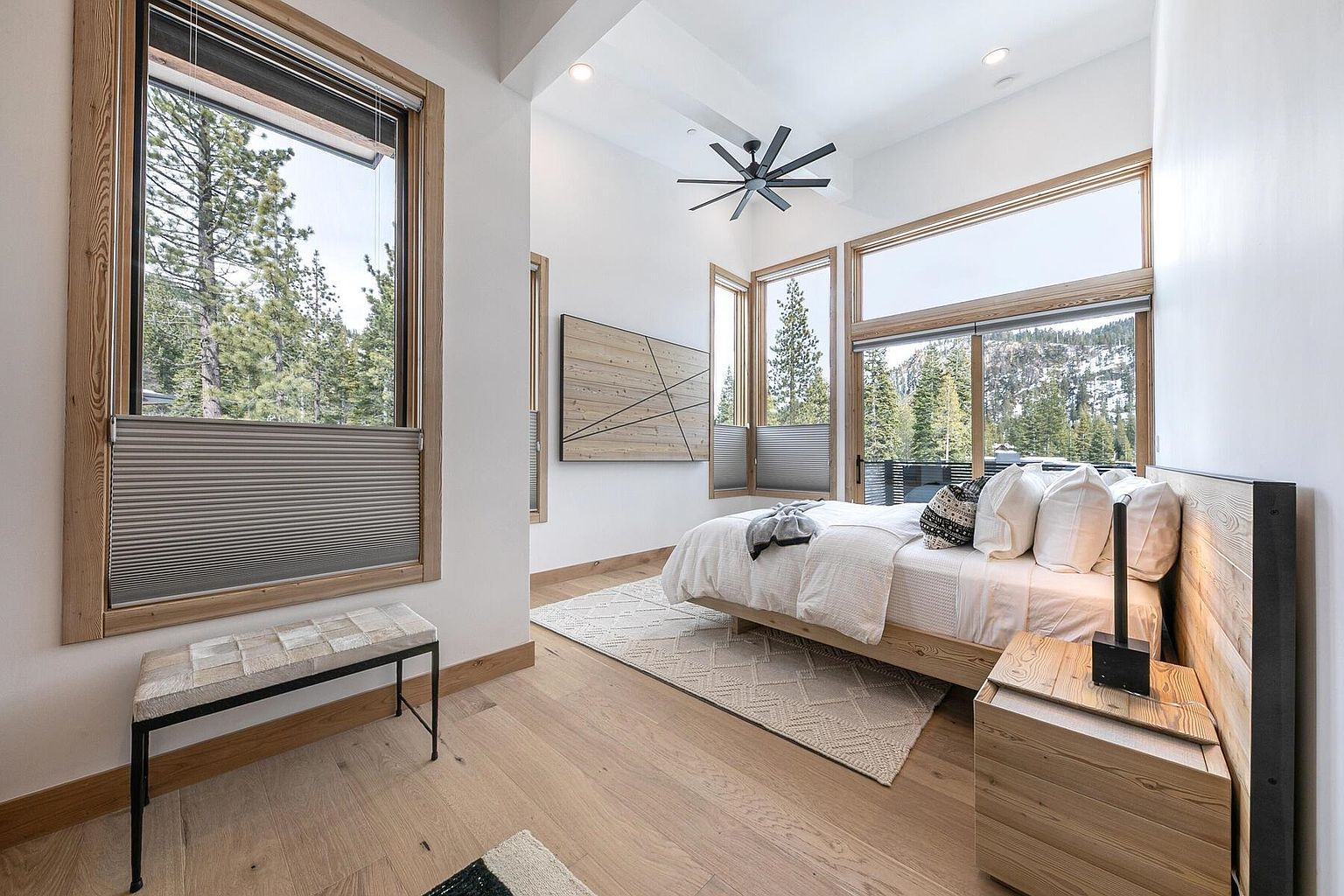 This is a bright and airy primary bedroom featuring large windows with natural wood frames that offer scenic views of the surrounding landscape. The room is decorated in a minimalist style with a light wood bed frame, white bedding, and a neutral-toned rug. A modern ceiling fan and a wooden accent wall add to the room's contemporary aesthetic.