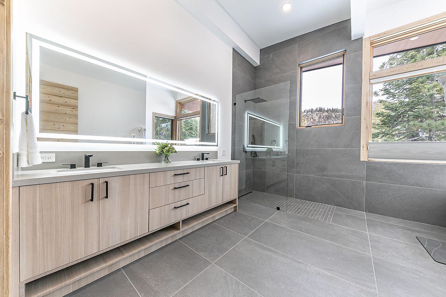 This is a modern primary bathroom featuring a double vanity with light wood cabinetry and black hardware, topped with a light-colored countertop. A large, illuminated mirror spans the length of the vanity. The bathroom also includes a glass-enclosed shower with gray tile and a window providing natural light and a view of the outdoors. The floor is covered in large gray tiles, contributing to the contemporary aesthetic.