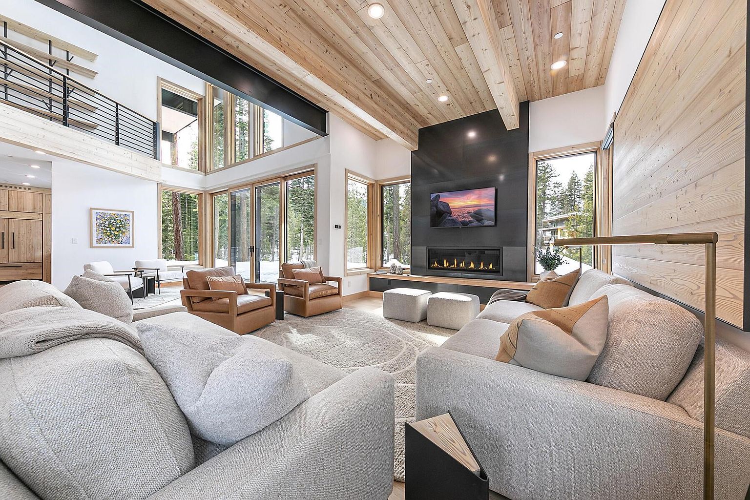 This is a spacious living room featuring a high ceiling with wooden planks, large windows offering natural light and views of the surrounding trees, and a modern fireplace with a mounted television. The room is furnished with comfortable sofas and armchairs, creating a cozy and inviting atmosphere. The overall style is contemporary with rustic elements.