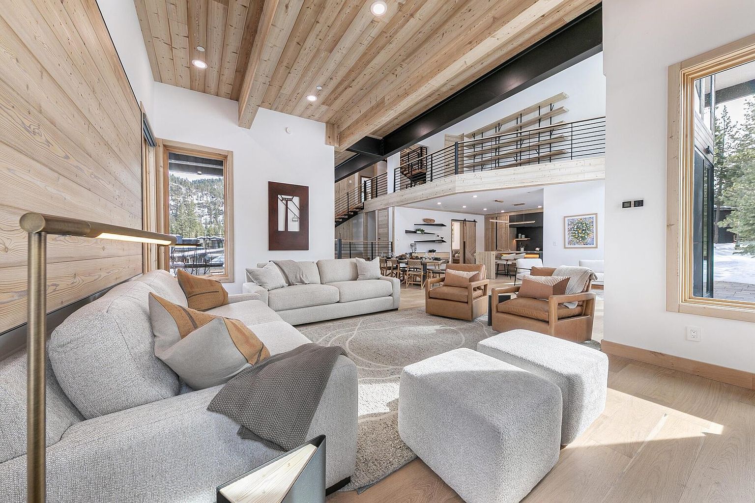 This is an interior shot of a modern living room with a high, wood-paneled ceiling and an open floor plan. The room features a large sectional sofa, two armchairs, and two ottomans, all arranged around a circular rug. A large window provides natural light and a view of the outdoors, while a staircase leads to a second-floor loft area.