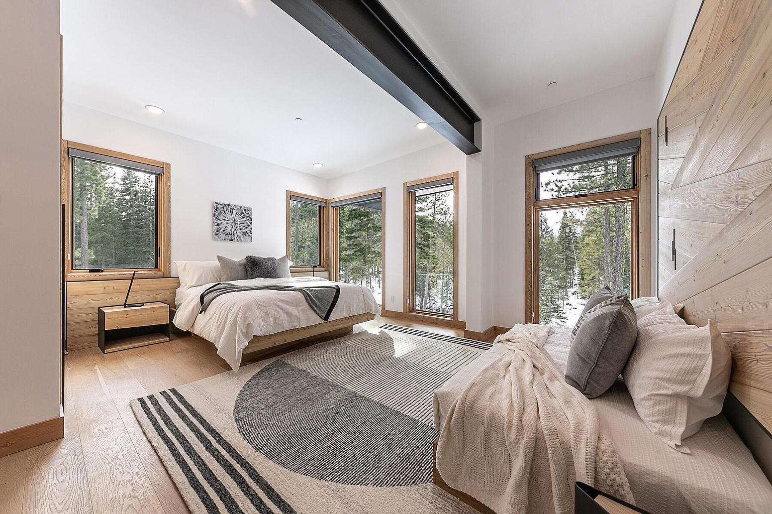 This is a bright and airy primary bedroom featuring a modern design with wood accents and large windows offering scenic views of the surrounding forest. The room includes a king-size bed with neutral bedding, a stylish area rug, and a cozy seating area, creating a serene and inviting atmosphere. The perspective is from the doorway, showcasing the entire room.