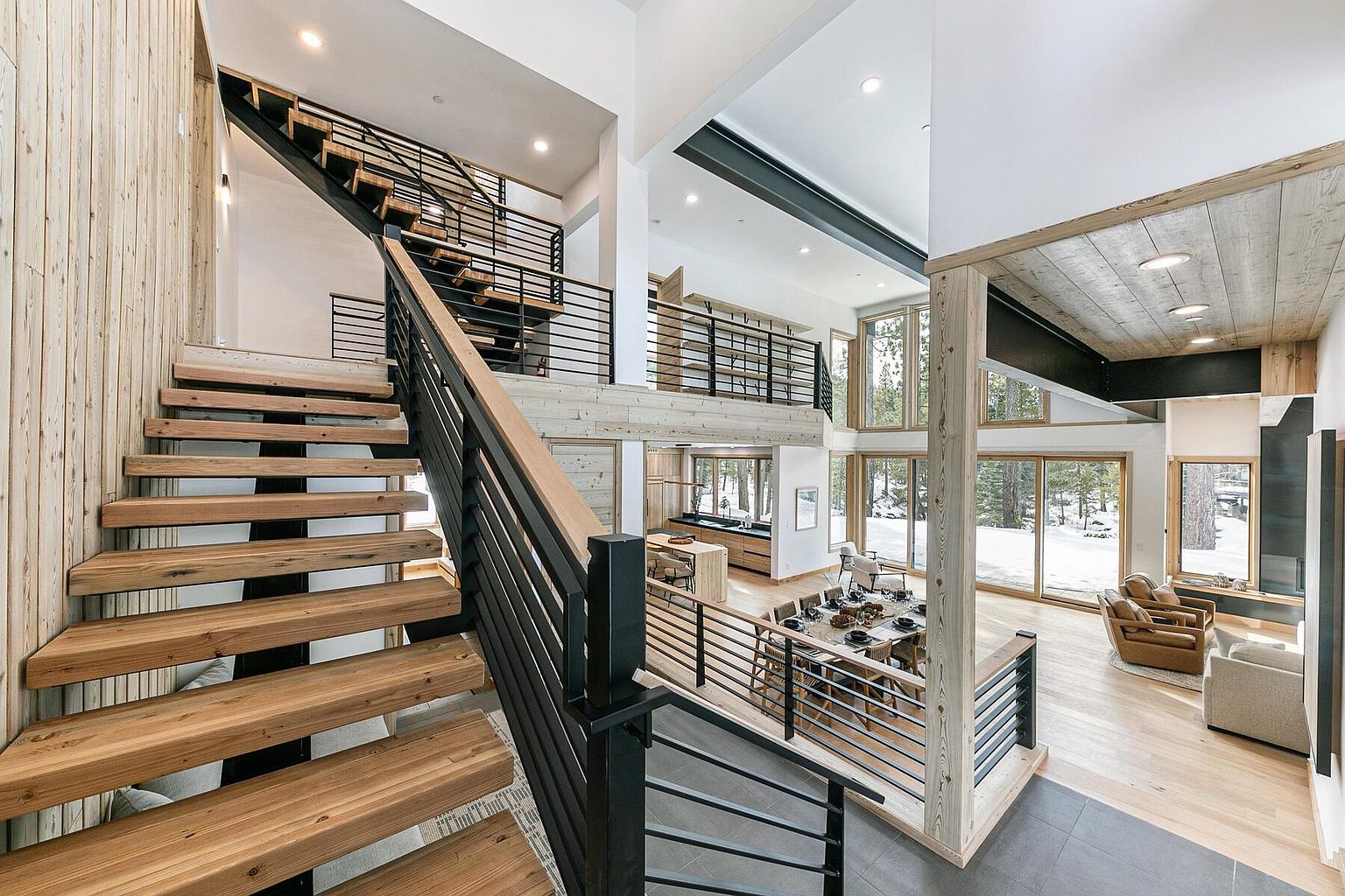 This interior shot showcases a modern home's grand hallway and staircase. The open-concept design features wooden stairs with black metal railings, complemented by light wood paneling on the walls. Large windows provide ample natural light and views of the snowy landscape, enhancing the home's connection to nature.