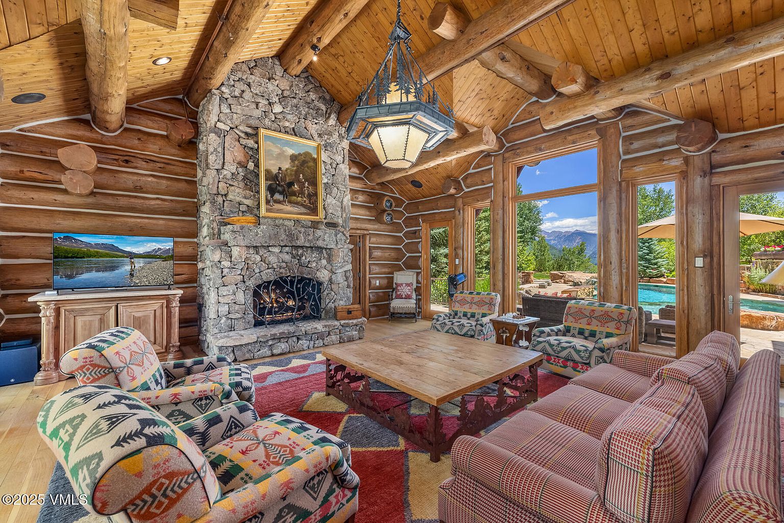 This is a warm and inviting living room featuring a rustic log cabin design with exposed wooden beams and a stone fireplace. The room is furnished with comfortable, patterned armchairs and a sofa, arranged around a wooden coffee table on a colorful rug. Large windows offer a view of the outdoor pool and landscape, creating a seamless indoor-outdoor living experience.