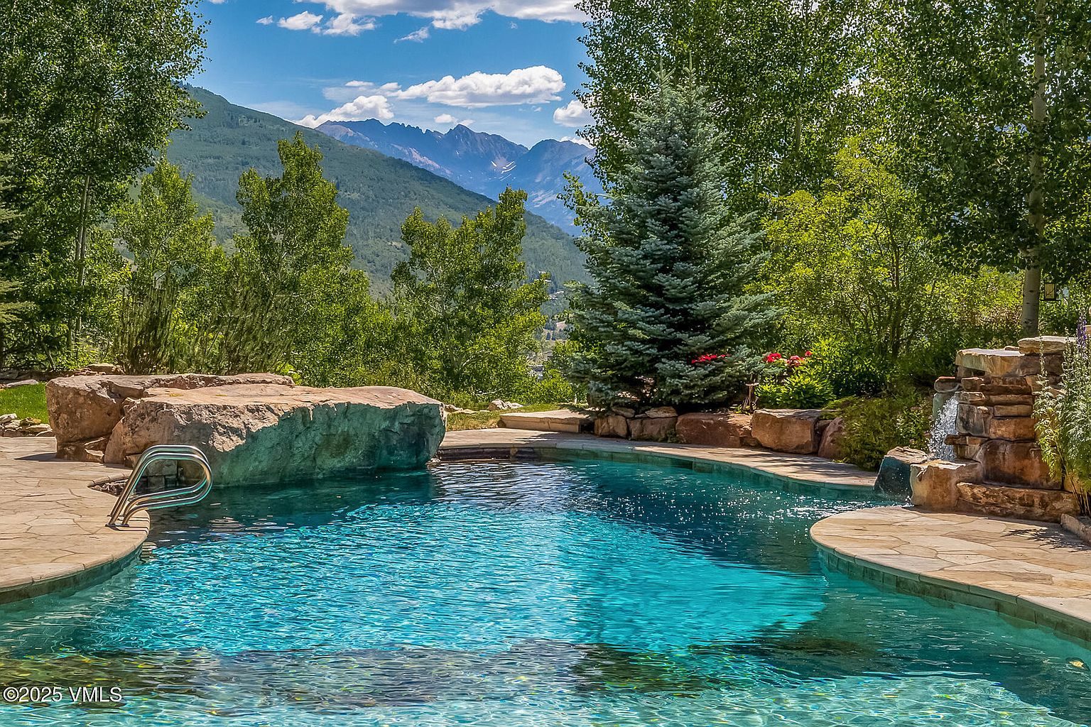 This image showcases a luxurious outdoor pool area with stunning mountain views. The pool features a natural stone surround, a small waterfall feature, and lush landscaping with trees and flowers. The clear blue water invites relaxation and enjoyment of the scenic surroundings, making it a desirable amenity for any property.