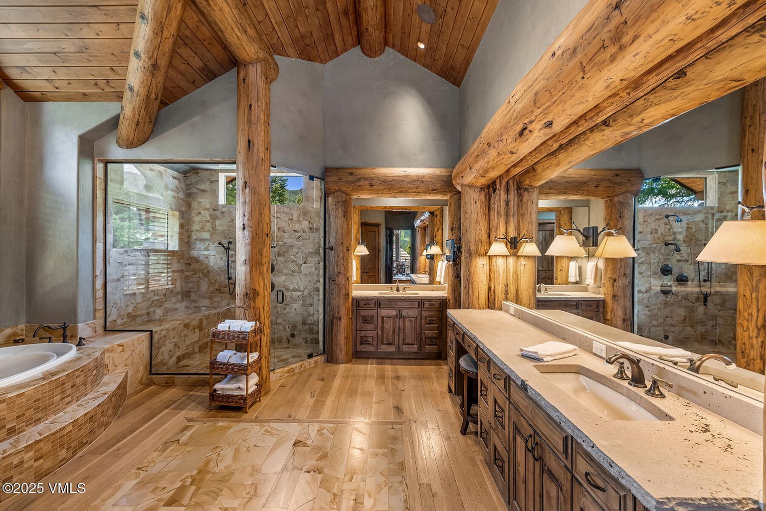 This is a luxurious primary bathroom featuring a rustic design with exposed wooden beams and log pillars. The bathroom includes a large vanity with dual sinks, a separate bathtub with mosaic tile surround, and a glass-enclosed shower. The flooring is a mix of wood and tile, and the overall impression is one of warmth and natural elegance.