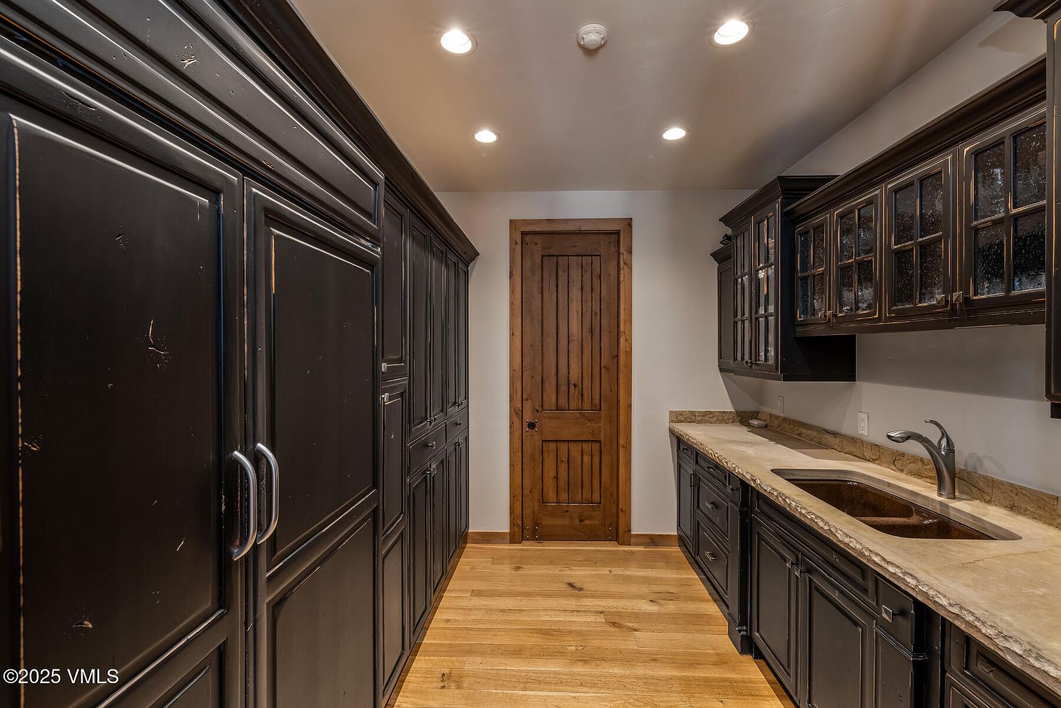 This is an interior shot of a pantry featuring dark wood cabinetry on both sides, providing ample storage space. A wooden door is visible at the end of the hallway, and the floor is made of light-colored hardwood. The pantry is well-lit with recessed lighting, creating a warm and inviting atmosphere.