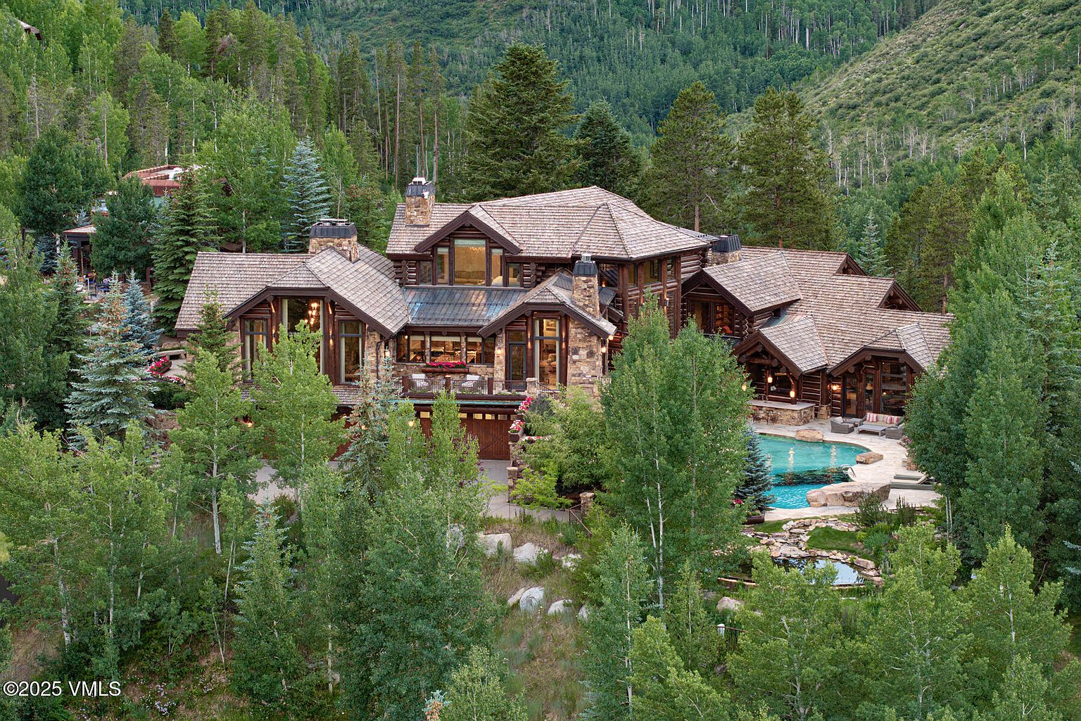 This aerial view showcases a luxurious log home nestled among lush greenery. The property features a multi-tiered roofline, stone chimneys, and a private pool area with a patio. The surrounding landscape is densely wooded, providing a sense of seclusion and natural beauty.