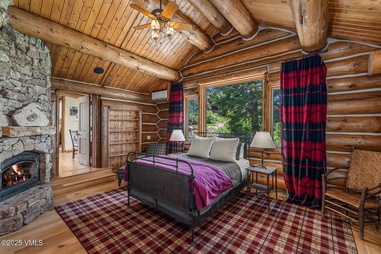 This is a cozy primary bedroom featuring a log cabin style with exposed wooden beams and walls. The room includes a stone fireplace, a wrought iron bed with a purple throw, and a window offering a view of the outdoors. The overall impression is rustic and inviting, perfect for a mountain retreat.