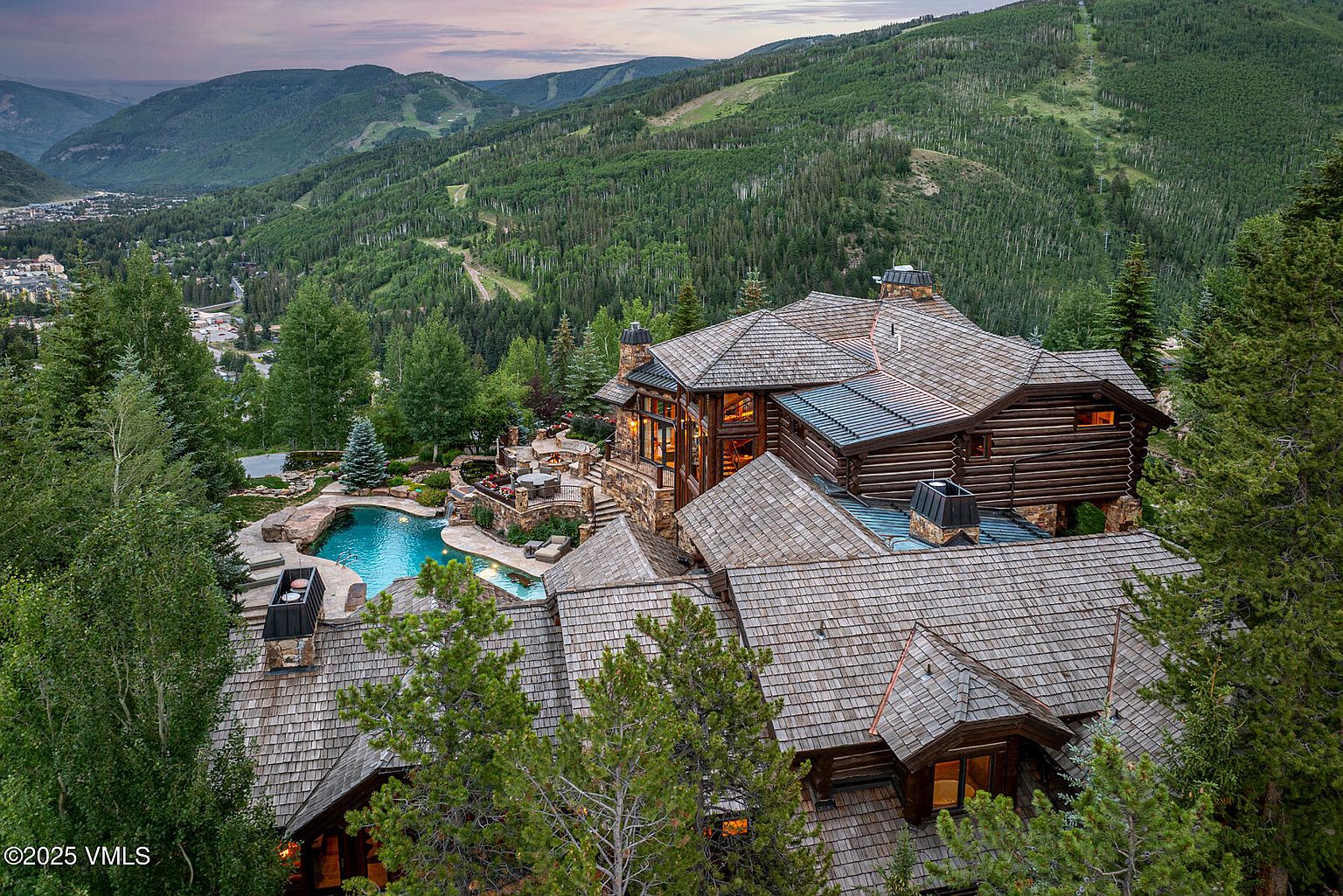 This aerial view showcases a luxurious log home nestled in a mountainous, wooded landscape. The property features a multi-tiered roof, a stone facade, and a stunning outdoor pool and patio area. The surrounding lush greenery and the home's architectural details create an impressive and inviting estate.