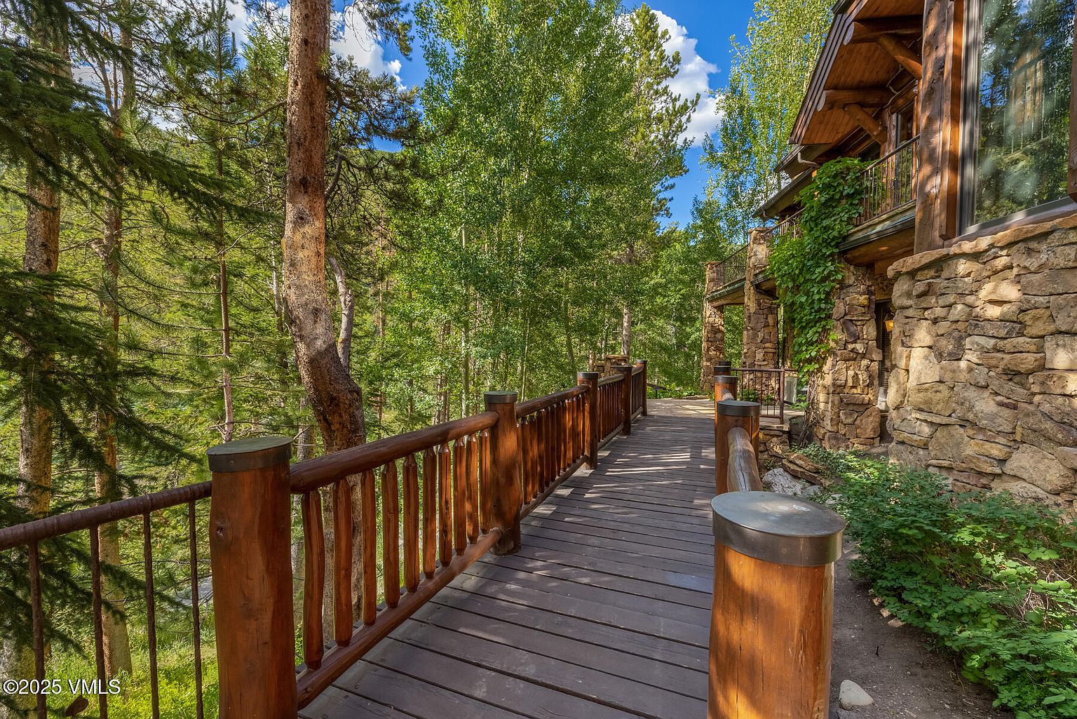 This image showcases a charming wooden walkway or deck, featuring a rustic wooden railing and posts. The walkway leads towards a stone-clad building with a balcony, suggesting a connection between the house and the surrounding natural environment. Lush greenery and trees create a serene and private atmosphere, enhancing the property's appeal.