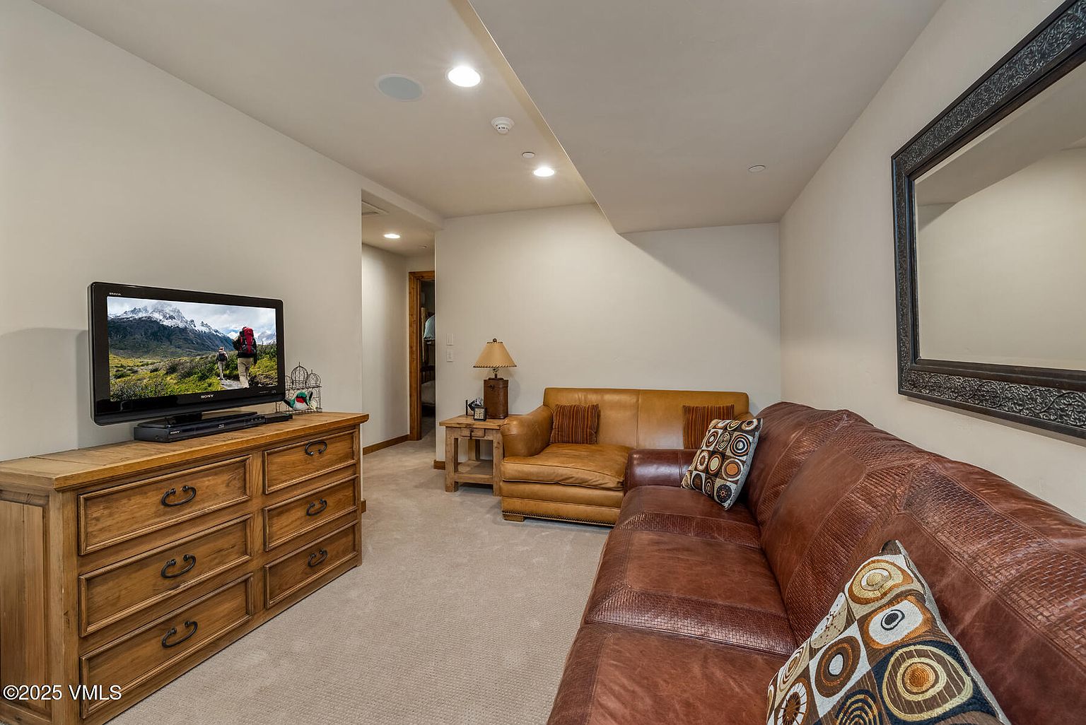 This is an interior shot of a cozy living room featuring a brown leather sectional sofa with decorative pillows, a wooden dresser with a television on top, and a framed mirror on the wall. The room is well-lit with recessed lighting and has a neutral-colored carpet, creating a warm and inviting atmosphere.