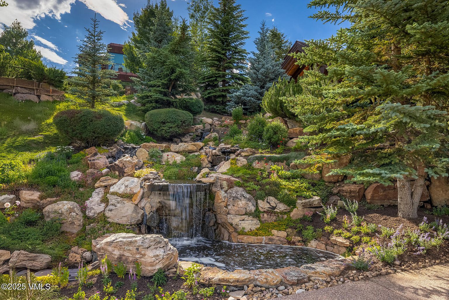 This image showcases a beautifully landscaped yard featuring a multi-tiered rock garden with a waterfall cascading into a small pond. Lush greenery, including various trees, bushes, and flowering plants, surrounds the water feature, creating a serene and natural ambiance. The scene is captured from a slightly low angle, emphasizing the height and depth of the garden.