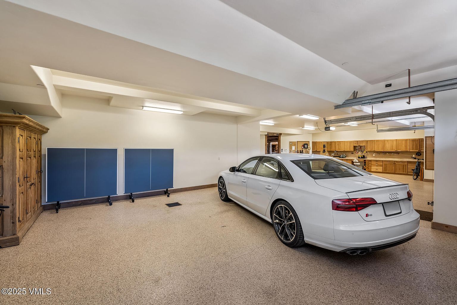 This is an interior shot of a spacious garage featuring a white Audi S8 parked on a speckled epoxy floor. The garage includes built-in wooden cabinets and countertops along the back wall, providing ample storage and workspace. A ping pong table is neatly folded and stored against the left wall, and the ceiling is well-lit with fluorescent fixtures.